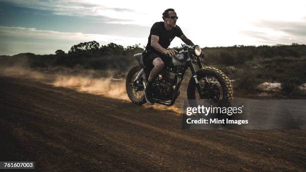 man wearing shorts and sunglasses riding cafe racer motorcycle on a dusty dirt road. - dirt road motorbike stock-fotos und bilder