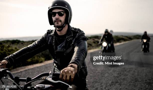 man wearing open face crash helmet and sunglasses riding cafe racer motorcycle along rural road. - biker fotografías e imágenes de stock