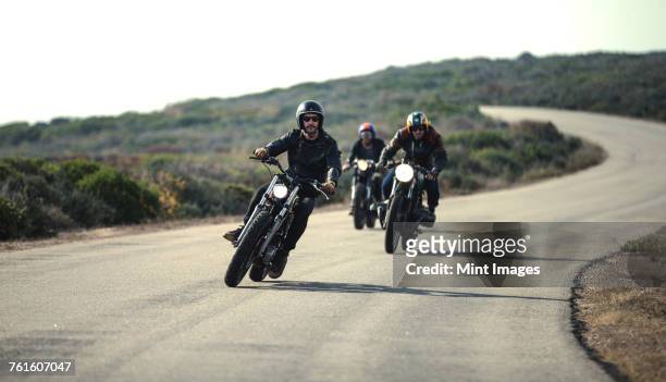 three men wearing open face crash helmets and sunglasses riding cafe racer motorcycles along rural road. - biker foto e immagini stock
