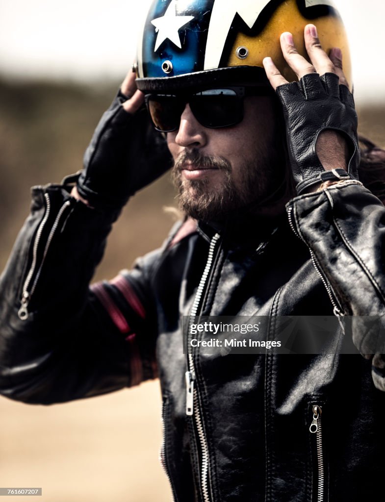 Bearded man wearing black leather jacket and sunglasses adjusting his yellow open face crash helmet.