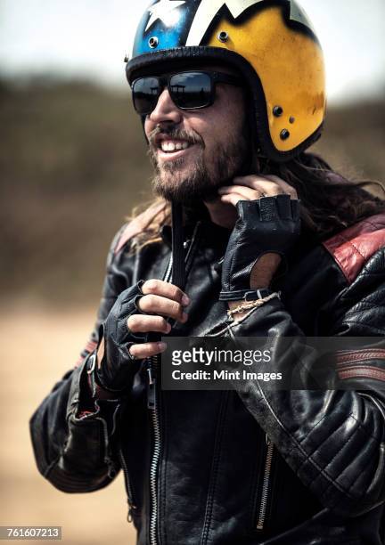 bearded man wearing black leather jacket and sunglasses adjusting his yellow open face crash helmet. - abrochar fotografías e imágenes de stock