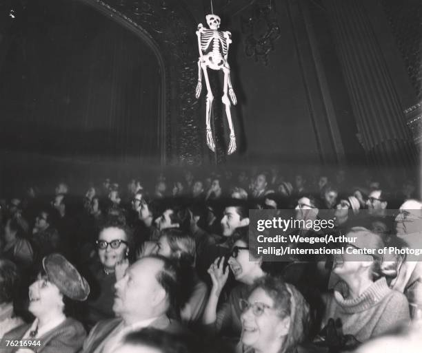 An audience laughs as an inflatable skeleton hangs over their heads, late 1950s or early 1960s. They are probably in a movie theatre during a screen...