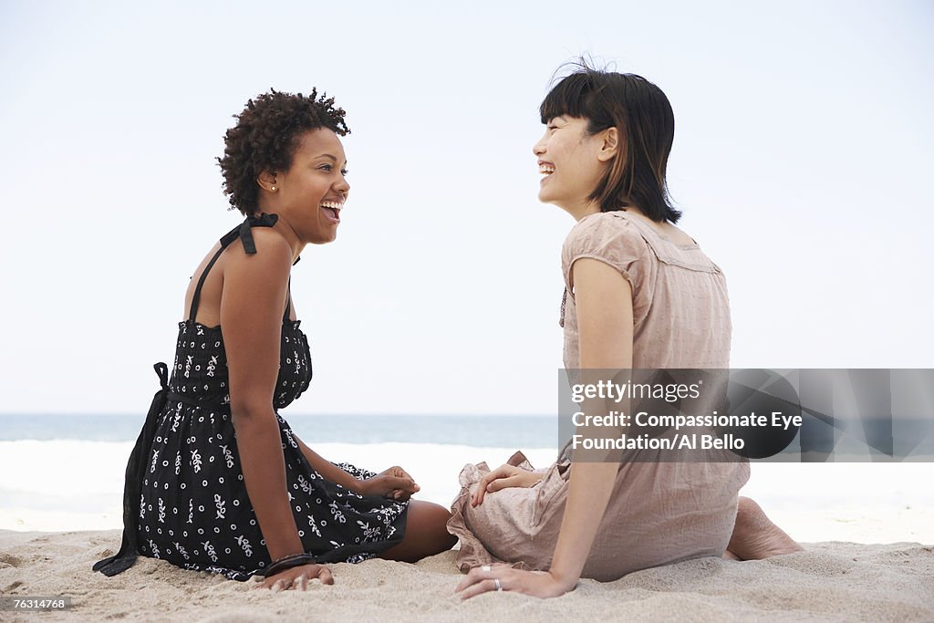 Two young women sitting on beach laughing