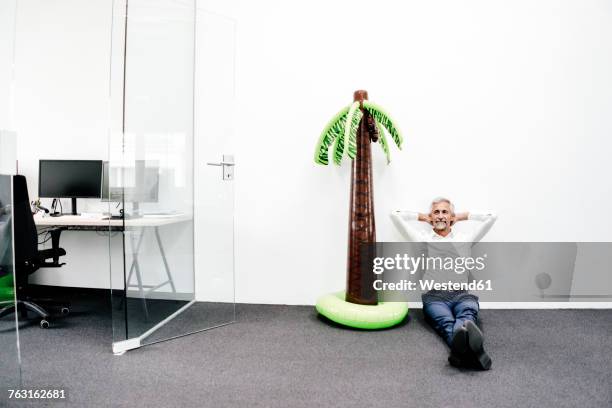 smiling mature businessman sitting next to inflatable palm tree in office - escapisme stockfoto's en -beelden