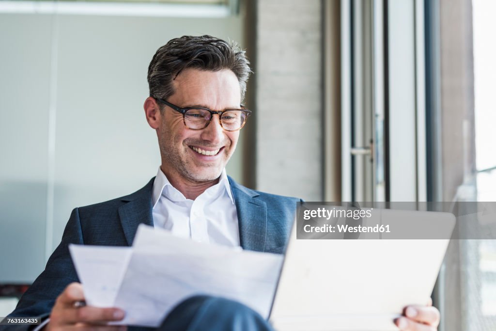 Portrait of laughing businessman with documents looking at tablet