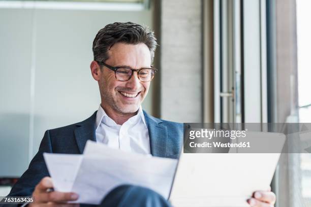 portrait of laughing businessman with documents looking at tablet - business man tablet stock pictures, royalty-free photos & images