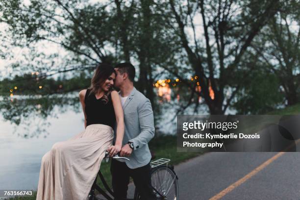 romantic young man whispering to girlfriend on bicycle handlebars by lake at dusk - ottawa park stock pictures, royalty-free photos & images