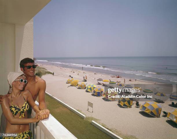 Smiling couple lean on a balcony at Thunderbird Motel, which overlooks a section of beach shaded by various colored umbrellas, Myrtle Beach, South...