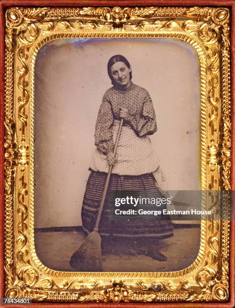 Gilt-framed tintype portrait of a woman in a polka-dot blouse and arpon over a striped skirt who holds a broom, mid 1860s.