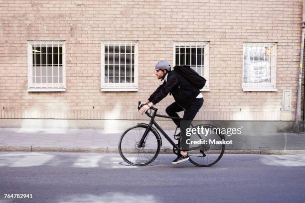 full length of businessman cycling on street by building in city - brick wall business person stock pictures, royalty-free photos & images