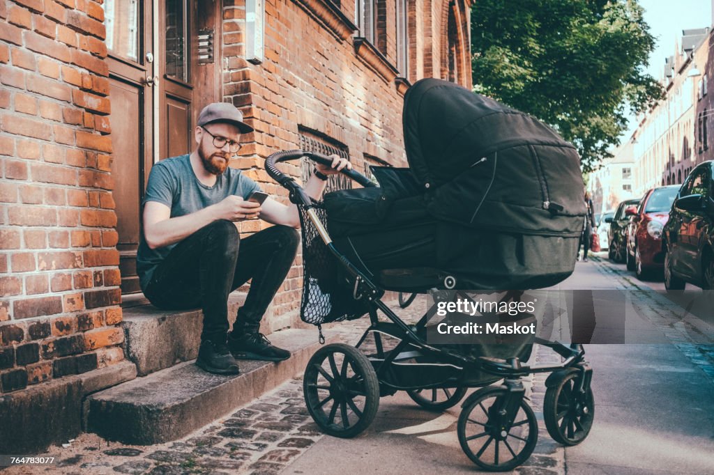 Father using smart phone while holding baby carriage on steps at building entrance