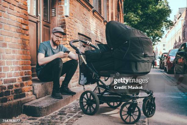 father using smart phone while holding baby carriage on steps at building entrance - baby stroller imagens e fotografias de stock