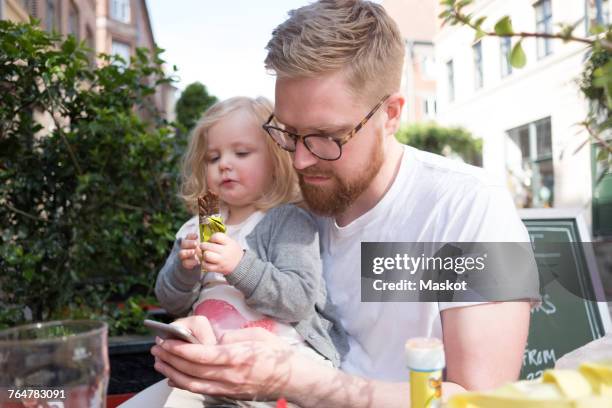 father using smart phone while sitting with daughter holding chocolate at sidewalk cafe - father and daughter looking at smartphone together stock pictures, royalty-free photos & images