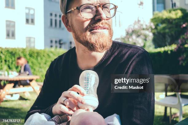 mid adult man looking away while feeding milk from bottle to baby boy at park - bébé biberon photos et images de collection