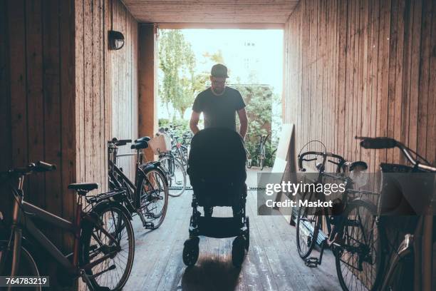 man pushing baby carriage while walking at corridor amidst bicycles and wooden walls - carriage stock pictures, royalty-free photos & images