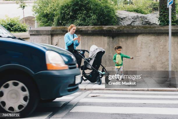 mother using mobile phone while standing with son and baby stroller on sidewalk at city - zebrastreifen stock-fotos und bilder