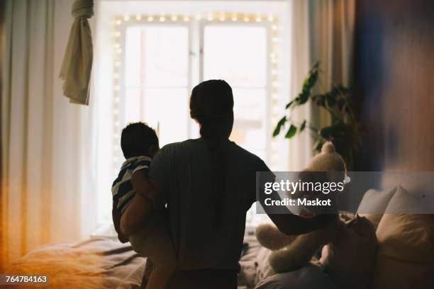 woman keeping teddy bear on bed while carrying son at bedroom - stereotypical housewife stock pictures, royalty-free photos & images