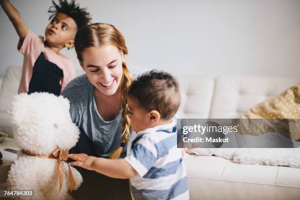 smiling mother showing teddy bear to toddler while boy playing by sofa at home - huisvrouw stockfoto's en -beelden