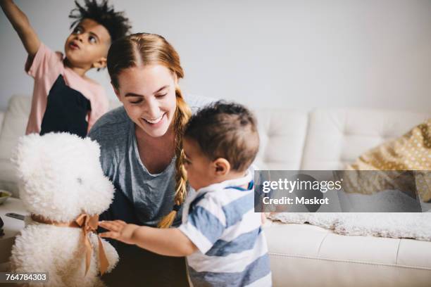 smiling mother showing teddy bear to toddler while boy playing by sofa at home - mama bear - fotografias e filmes do acervo