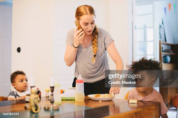 mother talking on mobile phone while standing by children at table - stereotypical housewife stock-fotos und bilder