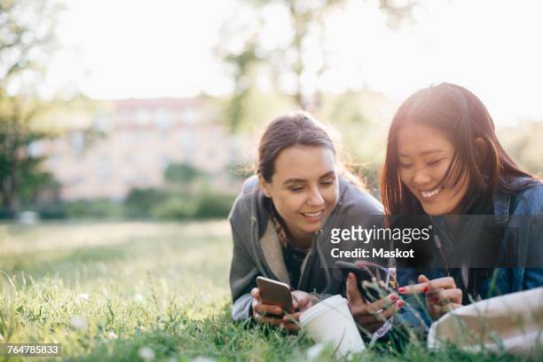 smiling female friends using smart phones while lying on grass - girl mobile stock-fotos und bilder