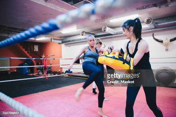 Female athlete kicking paddings held by woman in boxing ring at gym