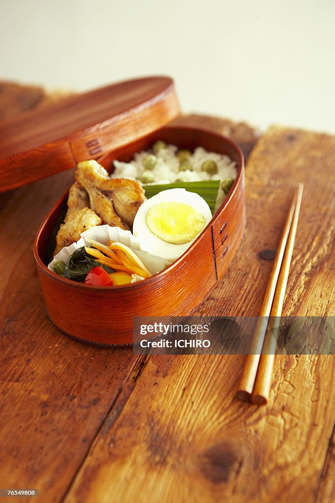 Japanese food in wooden lunch box, next to chop sticks on wooden table