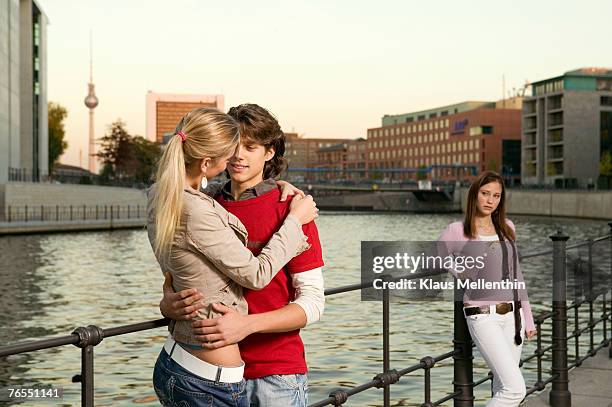 couple embracing at river, young woman standing in background - jaloezie negatieve emotie stockfoto's en -beelden