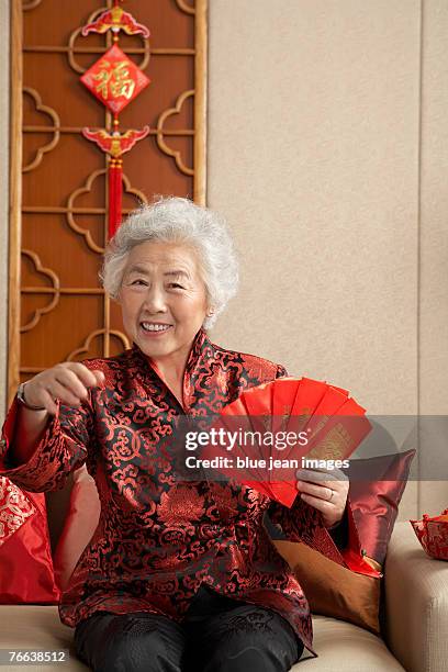 an old woman in chinese traditional clothing with red envelopes sitting on the sofa celebrates chinese new year. - 65 year old asian women ストックフォトと画像