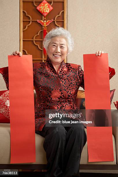 an old woman in chinese traditional clothing holding posters sitting on the sofa celebrates chinese new year. - 65 year old asian women ストックフォトと画像