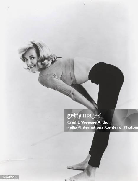 Portrait of American actress and model Chris Noel, barefoot and dressed in tight leggings and a cropped top shirt, as she bends at the waist, her...