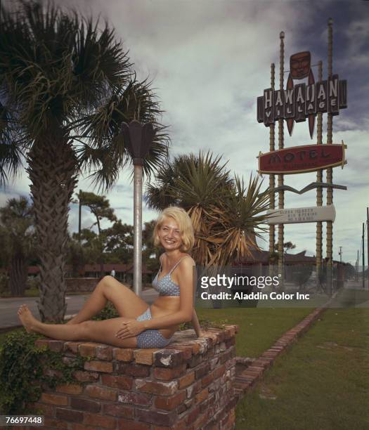 Woman in a bikini sits on a brick flower planter in front of the Hawaiian Village Motel, Myrtle Beach, South Carolina, 1960s.