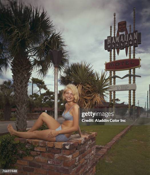 Woman in a bikini sits on a brick flower planter in front of the Hawaiian Village Motel, Myrtle Beach, South Carolina, 1960s.