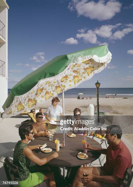 Casually dressed couples enjoy sandwiches and drinks in tall glasses as they sit under an umbrella at a round table on a patio near the beach behind...