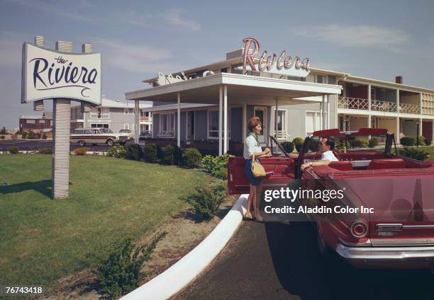 Woman talks with a man in the driver's of a convertible parked in the driveway in front of the Riviera motel, Bass River, Massachusetts, 1960s.