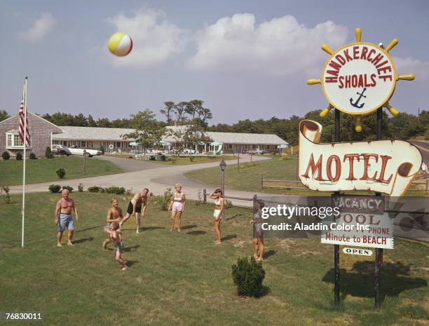 Group of people, adults and children, all dressed in swimwear play with a beach ball on the grass near the sign for the Hankerchief Shoals Motel,...