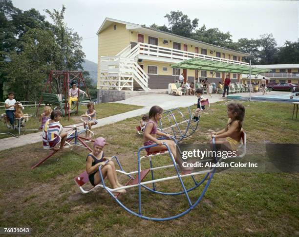 Children in swimwear play on several wire-frame see-saws on the grass at the Jolly House Motel & Resort, in the Catskills, New York, 1960s. A girl in...