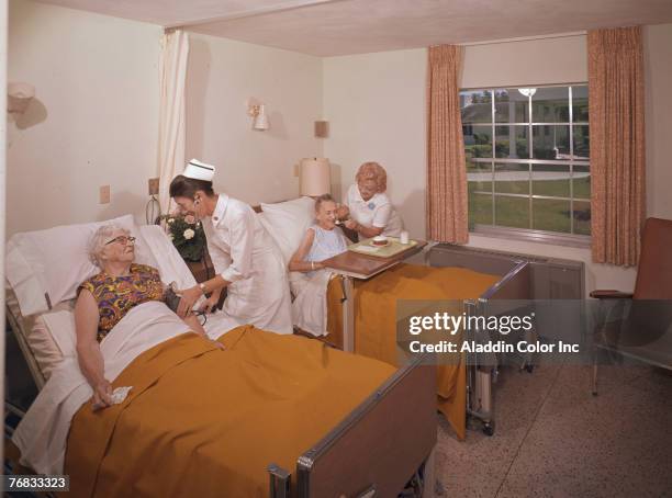 Pair of nurses work with two elderly women in a double occupancy room in an unidentified medical facility, 1960s. The nurse at right checks the blood...