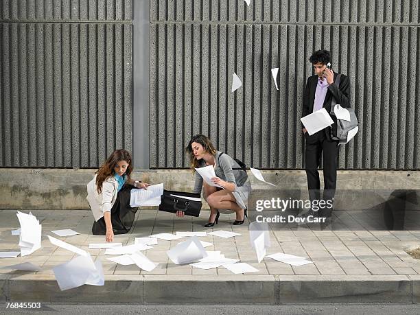 two businesswomen picking up papers blowing about pavement while a young man looks on, talking on the phone, - egoismus stock-fotos und bilder