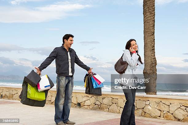 woman using mobile phone, man holding out shopping bags in background - selfishness stock pictures, royalty-free photos & images