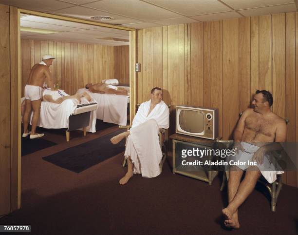 In a wood-panneled spa, two men in towels sit on either side of a television set and talk at a spa, 1960s. In the room beyond them, a two men, also...