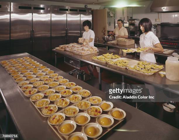Three uniformed women prepare food for serving trays in an unidentified institutional kitchen, 1960s. One arranges plastic-bagged sandwiches, a...