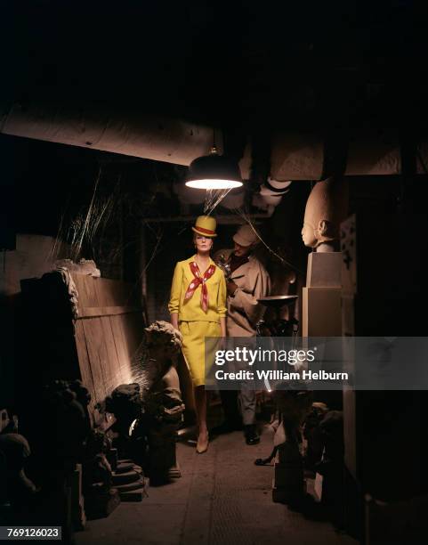 American model Angela Howard, in a mustard yellow skirt suit and matching derby hat, poses while an unidentified male model, with a pipe in his mouth...