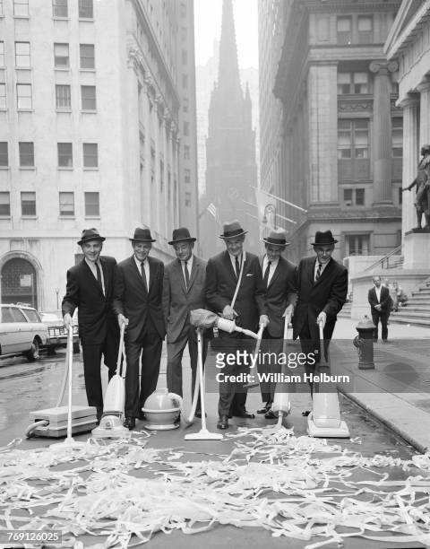 View of six unidentified men, all dressed in hats, suits, and ties, as they use a variety of different styles of vacuum cleaner to clear ticker tape...