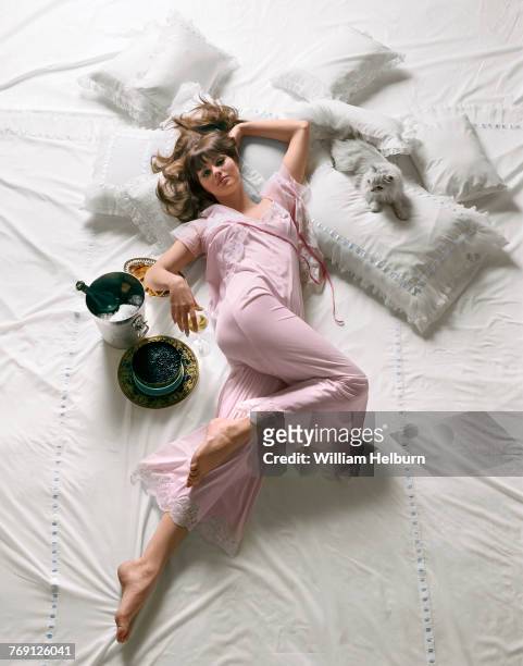Portrait, looking from above, of American model Sandra Hilton, dressed in pink pajamas and holding a wine glass in one hand, as she lies amid white...