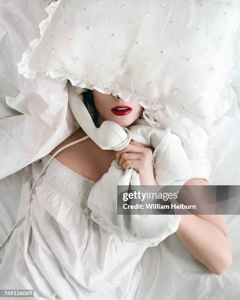 View of American actress and model Suzy Parker , in a white nightdress, as she lies on her back, a pillow over her eyes, and a white telephone...