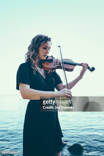beautiful young woman playing violin while standing by lake against clear sky - young woman standing against clear sky ストックフォトと画像