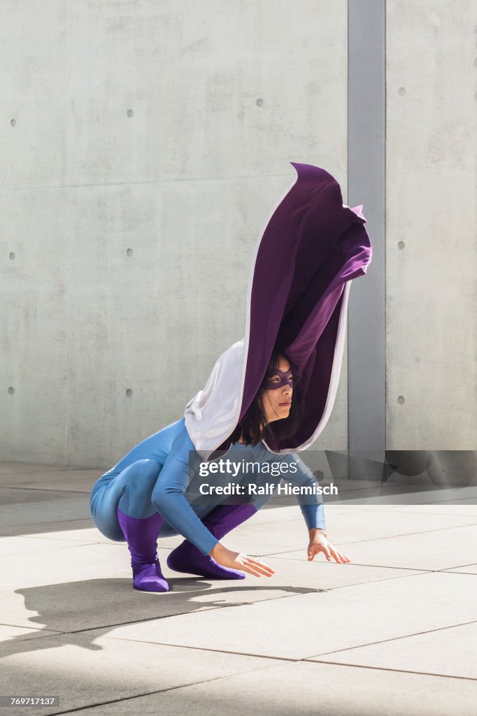 Female superhero wearing purple cape crouching on floor by wall