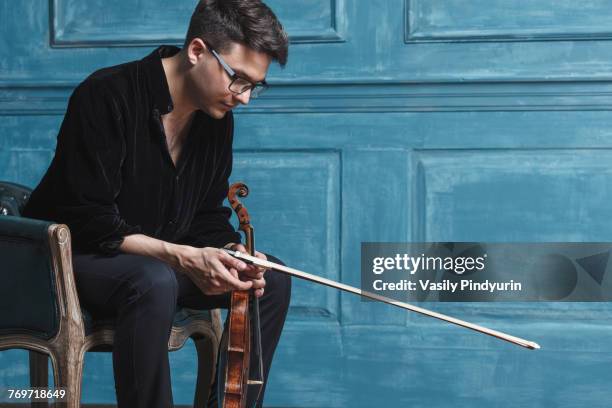 young man holding violin while sitting on chair by blue wall - violin scroll stock pictures, royalty-free photos & images