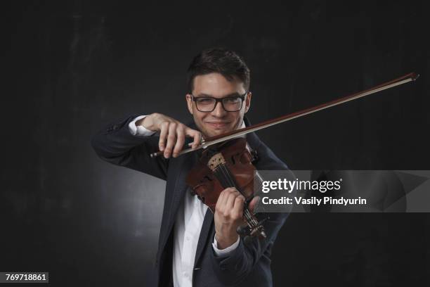 confident smiling young man playing violin while standing against black background - violin scroll stock pictures, royalty-free photos & images